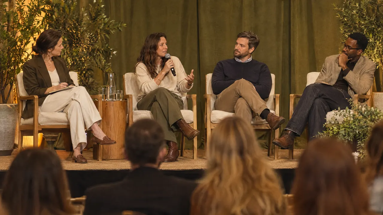 Four panelists on a conference stage listen as one of them speaks into a microphone.